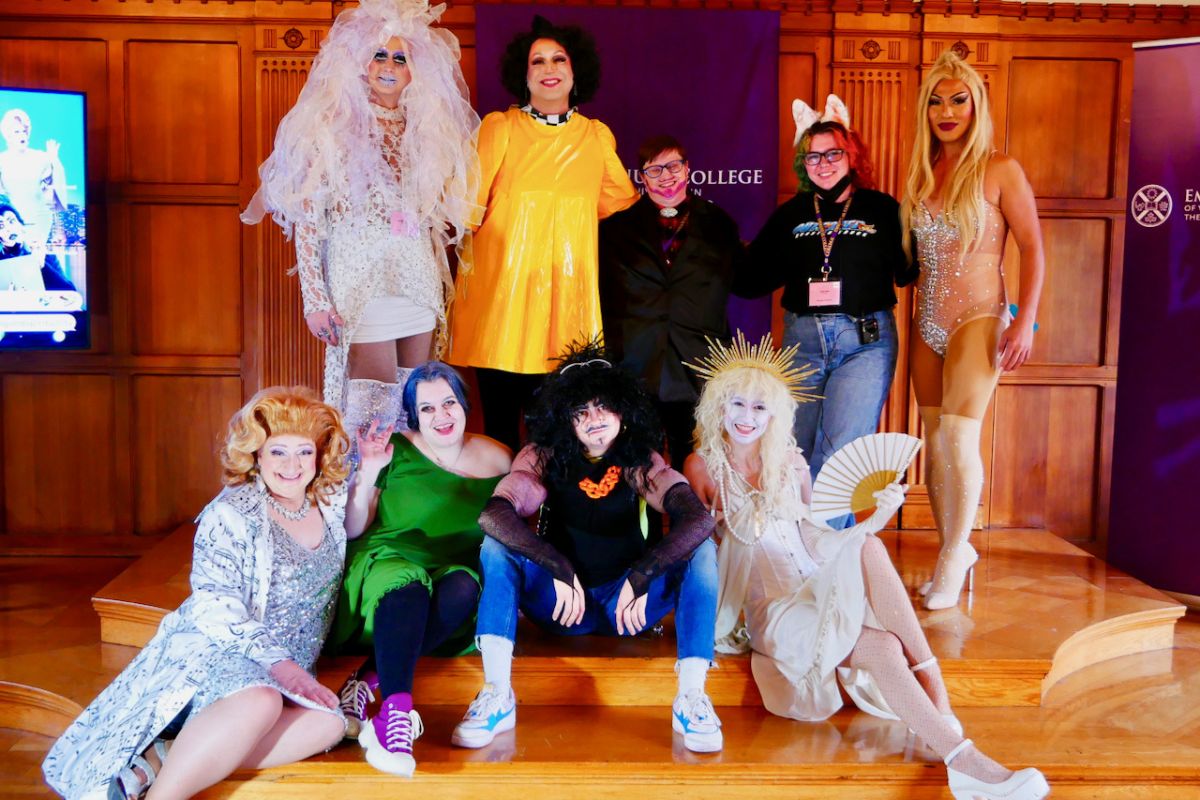 A group of performers in drag stay standing or sitting at the front of a room.