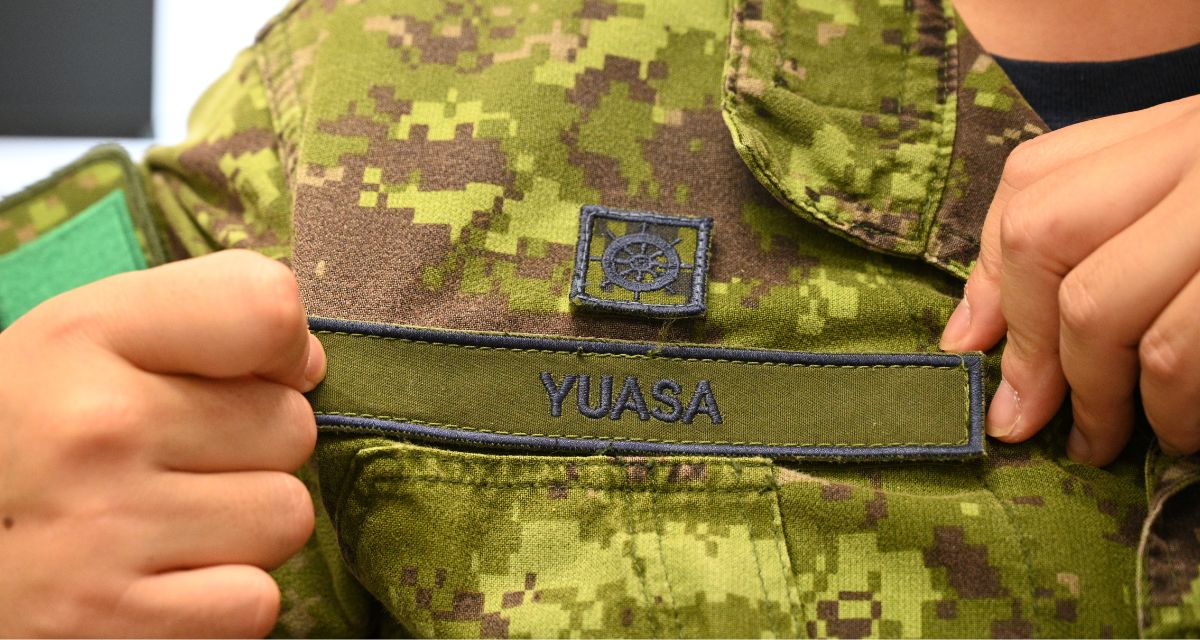 Hands are positioned to highlight the name Yuasa on a name badge attached to a set of green Canadian Forces army fatigues.