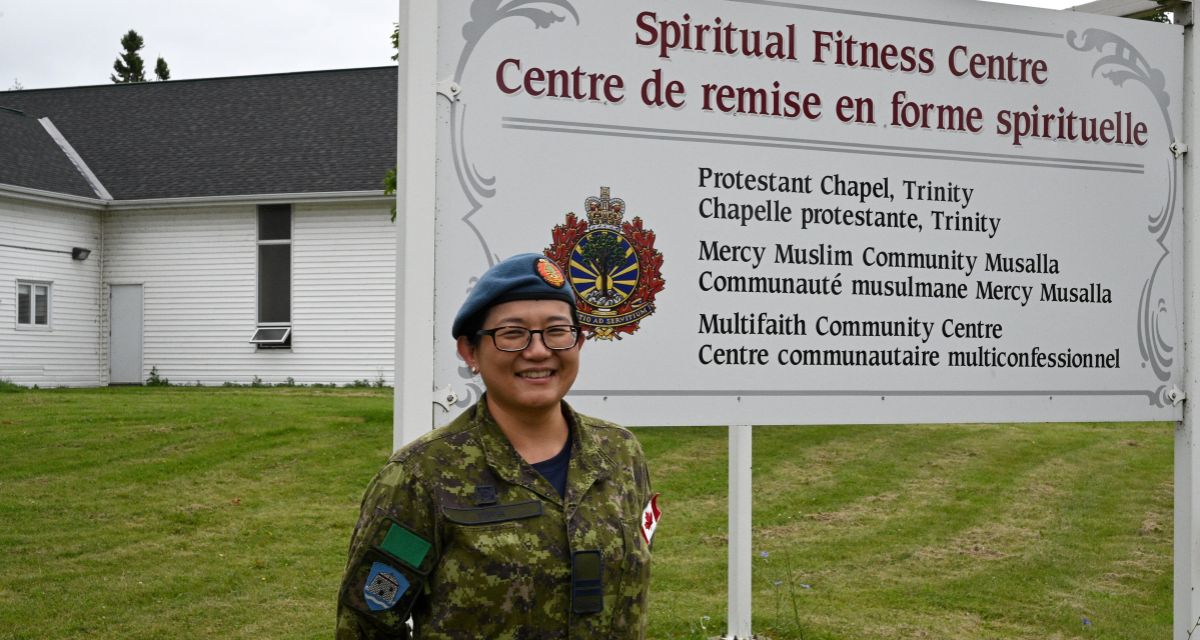 Capt. Yuasa stands in front of a sign reading Spiritual Fitness Centre in Kingston. Behind her, there is a lush green lawn and a white building that forms the backdrop of the scene.